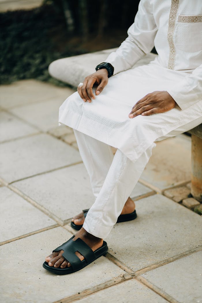 A man wearing traditional white attire and sandals sitting on a stone bench outdoors.