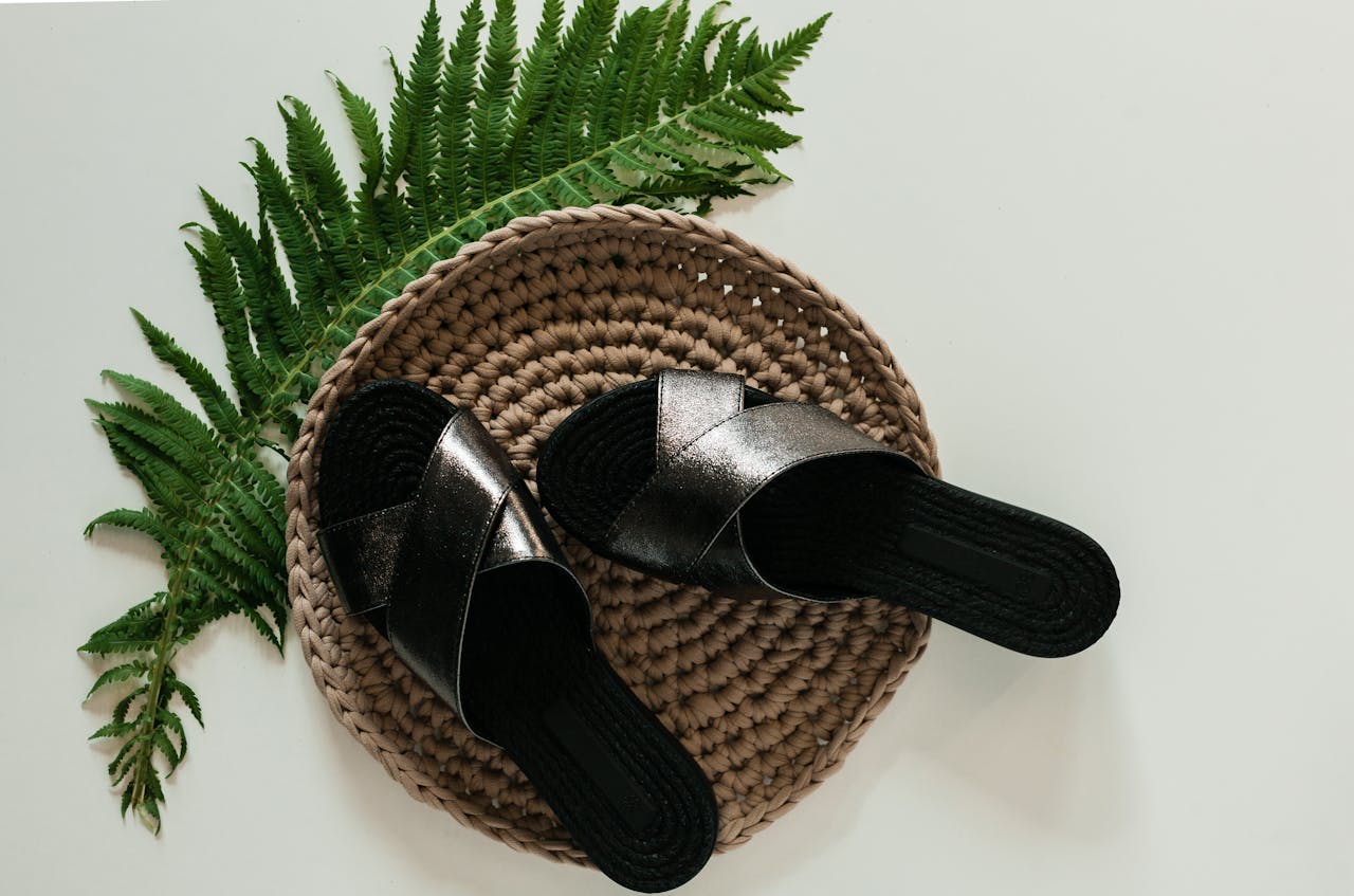 Pair of elegant black sandals placed on a woven mat with a decorative fern leaf.